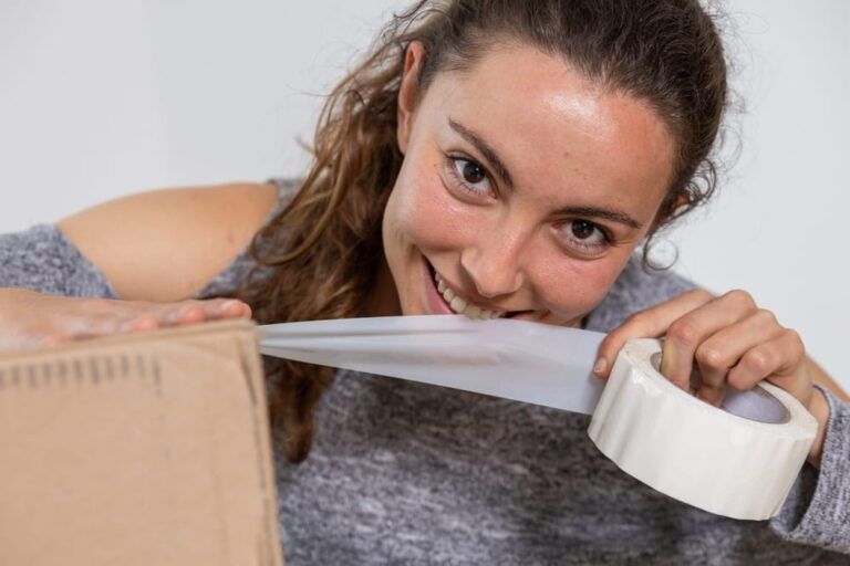 Happy pretty young woman packing boxes to move to her new home, cutting packing tape with teeth